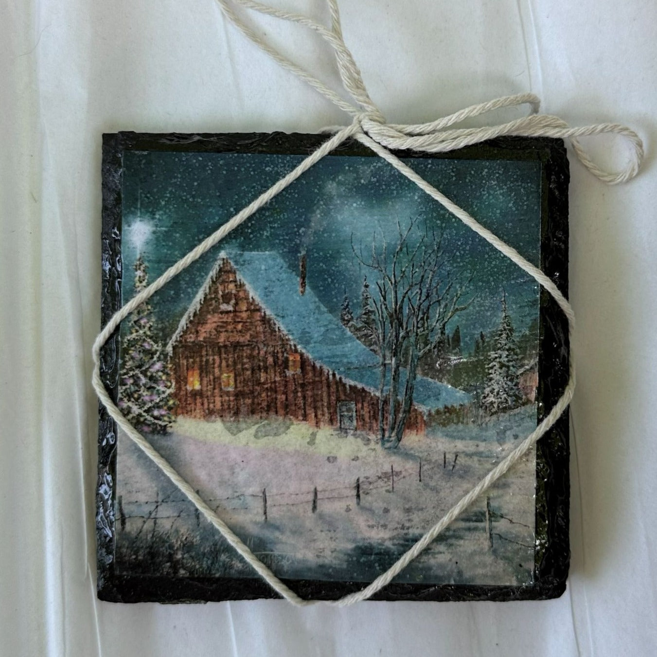 Decorative tile with a winter scene of a cabin, Christmas trees, and snow, tied with twine on a white background.