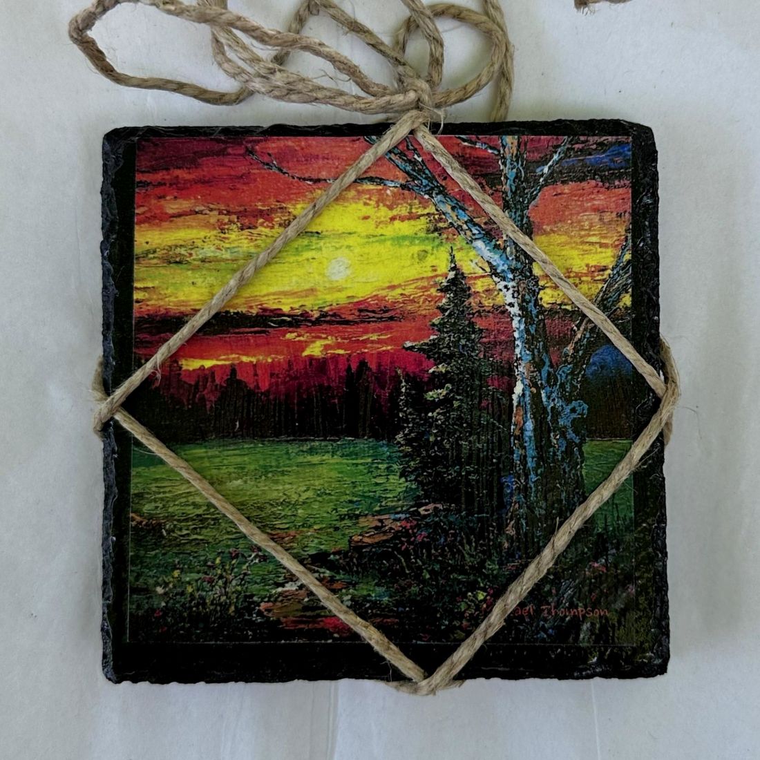 Square slate with a painted sunset scene and tree, tied with twine on a white background