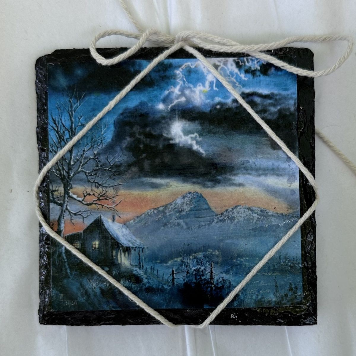 Decorative slate with a painted night scene of a house and mountains, tied with twine.