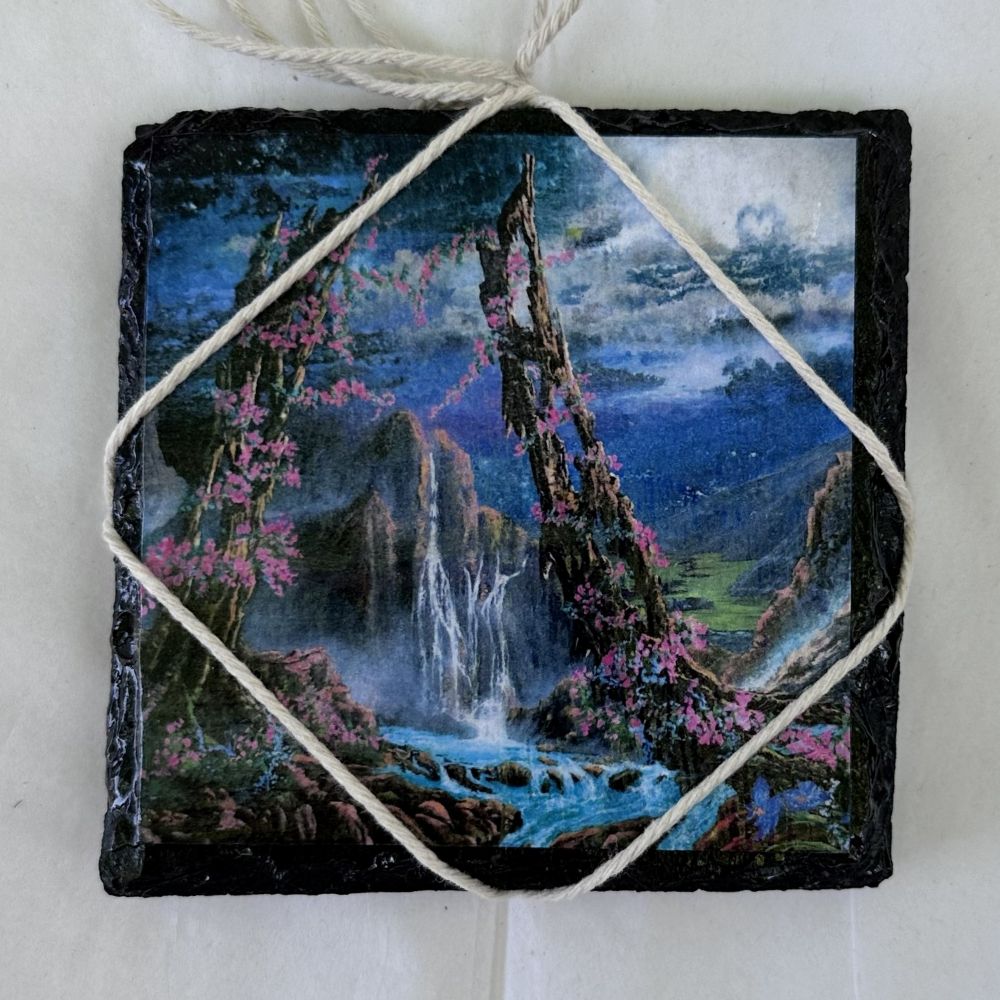 square slate coaster with a mountain and waterfall design, tied with twine on a white background.