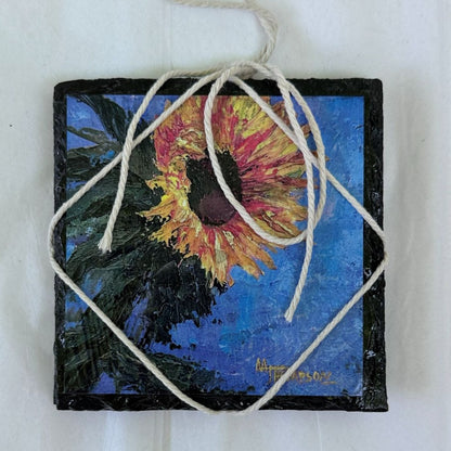 Square painting of a sunflower with a blue background, hanging by a string.